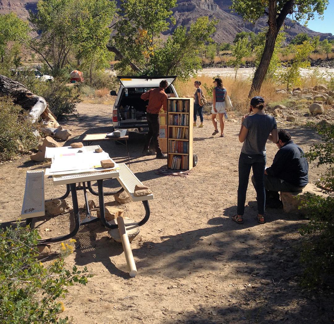A library and readers sit on a picnic table and the back of a pickup truck next to a river in a redrock canyon.