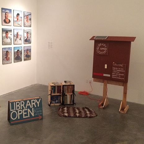 A library bookshelf, open sign, and interpretive sign sit in a gallery.